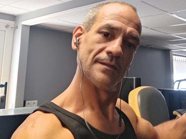 Primer plano de FairlightFrancais, un hombre calvo de mediana edad con una ligera sonrisa, que lleva una camiseta sin mangas negra y auriculares. Está sentado en un escritorio, sosteniendo un auricular amarillo. Su mirada es increíblemente intensa..