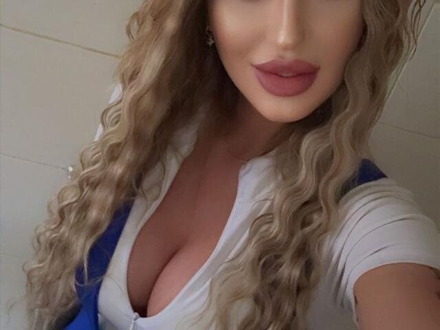 NicolSNaya with curly blonde hair, wearing a low-cut white shirt, showing large boobs, pink lipstick, and a blue backpack, taking a selfie.