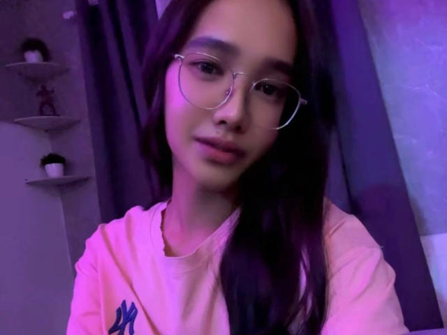 Close-up photo of SooHotN2Sexy-hot, a young woman with long brown hair, wearing clear glasses and a pink hoodie. Soft purple lighting, slightly seductive expression, visible shelves in the background.