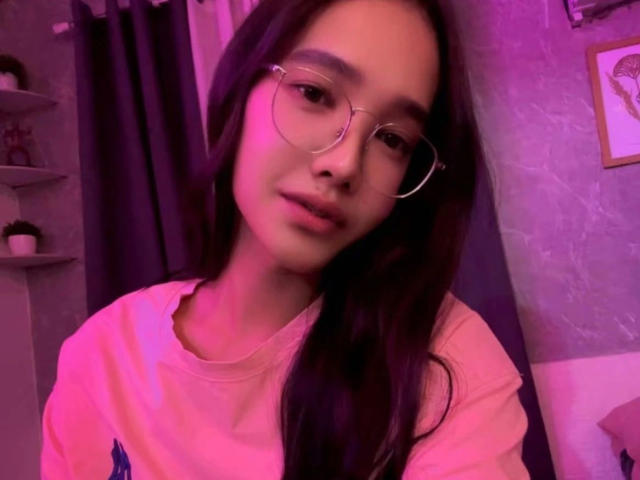 Gros plan de SooHotN2Sexy, une femme séduisante avec de longs cheveux noirs, portant des lunettes rondes et une chemise rose. L'éclairage violet projette une lueur séduisante sur son visage, créant une forte tension sexuelle. Expression douce.