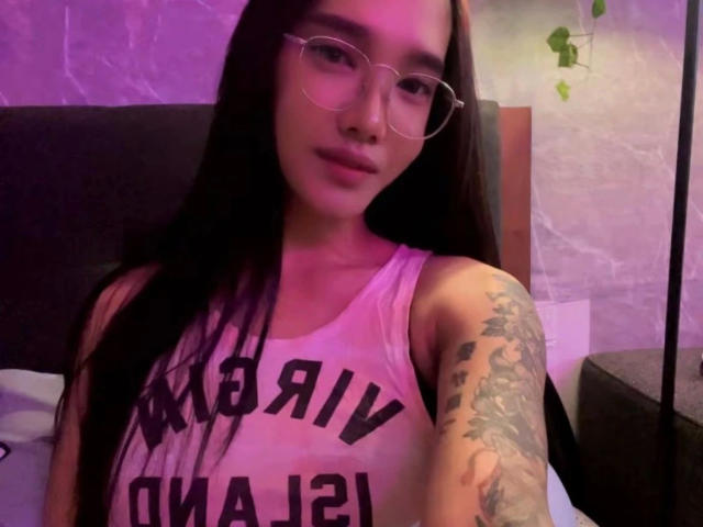 SooHotN2Sexy - muito atraente, com cabelos longos e pretos, vestindo uma camiseta branca com letras pretas, óculos de grau e um braço tatuado. Olhando diretamente para a câmera com uma expressão sexy e sedutora. Iluminação roxa.