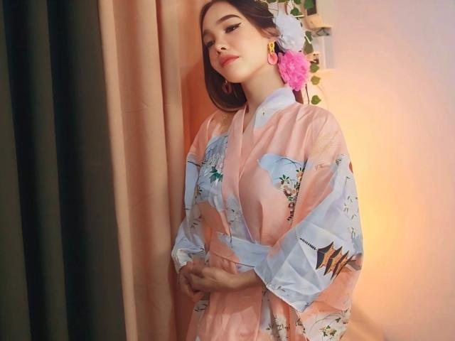 SooHotN2Sexy: aantrekkelijk, met halflang bruin haar, draagt een doorschijnende roze kimono met bloemenmotieven, en staat voor een bruine achtergrond. gordijn. Roze bloem in het haar, gouden oorbellen, zelfverzekerde pose, sensuele spanning..