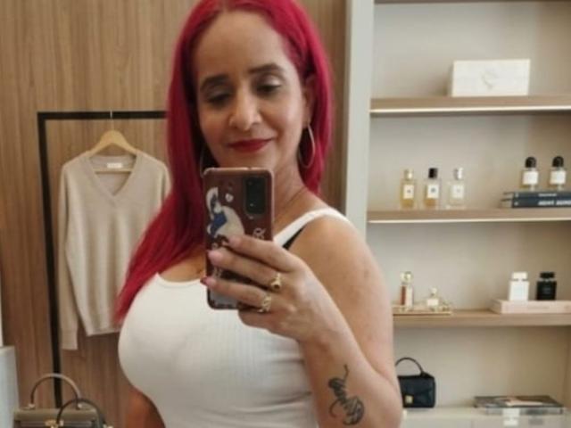 KateMonroe, avec ses cheveux roux éclatants, prend un selfie devant un miroir, tenant un téléphone avec une coque noir et blanc. Elle porte un débardeur blanc moulant qui met en valeur sa poitrine généreuse. Tatouage sur l'avant-bras gauche. Étagères avec des flacons de parfum en arrière-plan. Avec un sourire en coin.