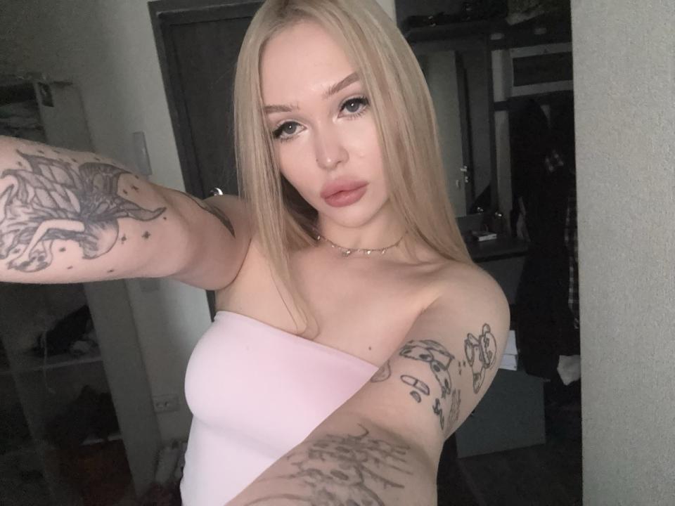 HoneyDripX, avec de longs cheveux blonds, une peau pâle et des tatouages sur les deux bras, porte un haut rose sans bretelles. Elle prend un autoportrait, légèrement incliné, avec un regard séduisant. Fond légèrement éclairé.