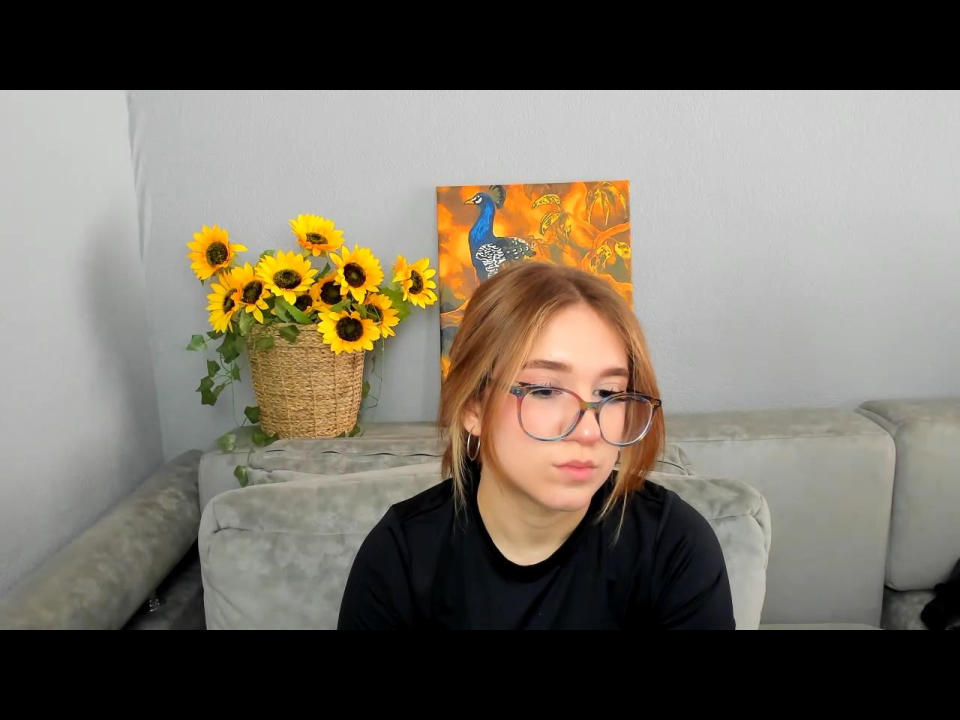 FellDivain, with red hair and glasses, sits on a gray couch, wearing a black shirt, next to a basket of sunflowers and an orange painting. She's looking directly at the camera, with a slightly aroused expression.