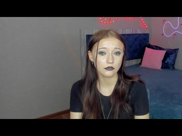 OliviaGilson has pale skin, dark brown hair, and black lipstick. She wears a black shirt and a necklace. Red string lights and neon outlines create a sexual tension background.