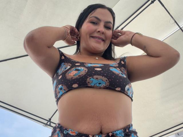 Scarlett Oxford, eine kurvige lateinamerikanische Frau mit langen, schwarzen Haaren, trägt ein schwarzes Crop-Top mit bunten Mustern, die Hände hinter dem Rücken. ihr Kopf, lächelnd in die Kamera, wobei ihr Bauch und ihre Taille sichtbar sind..