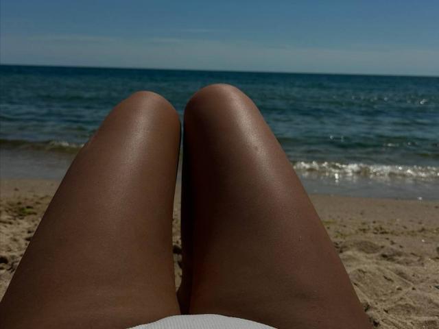 Fotografie von Elionorias dunkelbraunen, glänzenden Beinen, die auf einem sandigen Strand auseinander gespreizt sind, mit dem blauen Ozean und dem klaren Himmel. im Hintergrund. Elionorias Beine bilden einen auffälligen, erotischen Rahmen..