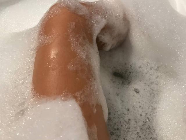 Fotografia em close-up da perna e coxa de Elionoria, parcialmente submersas em água com sabão. Bolhas espumosas cobrem a pele, realçando o brilho de sua pele bronzeada. A perna está dobrada no joelho..