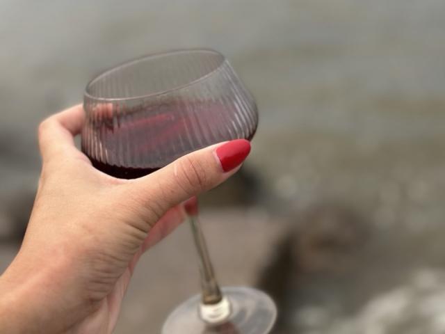 Close-up de uma mão com unhas pintadas de vermelho segurando um copo de vinho tinto escuro. A mão está ligeiramente inclinada, mostrando a curvatura do copo. O fundo é água desfocada..