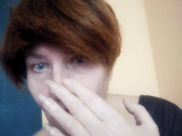 Photo en gros plan de MioritaStar, avec des cheveux bruns courts, des yeux bleus et une peau pâle. La main de MioritaStar recouvre sa bouche, et elle regarde directement la caméra avec une expression intense et légèrement nerveuse..