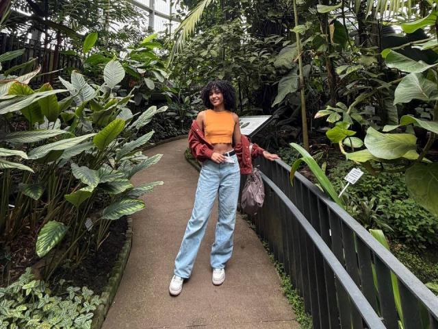 CinthiaWalker se tient sur un sentier étroit et luxuriant, entourée d'une végétation dense. Elle porte un haut orange vif, une veste rouge à motifs, et un jean bleu clair, avec des baskets blanches. Elle se penche contre une rambarde noire.