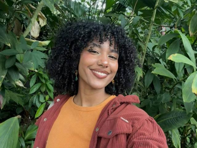 Cinthia Walker, avec une peau brun clair et des cheveux noirs bouclés, souriant largement. Portant un pull orange et une veste en velours rouge, debout devant un feuillage vert luxuriant. La lumière vive du jour met en valeur son expression joyeuse..