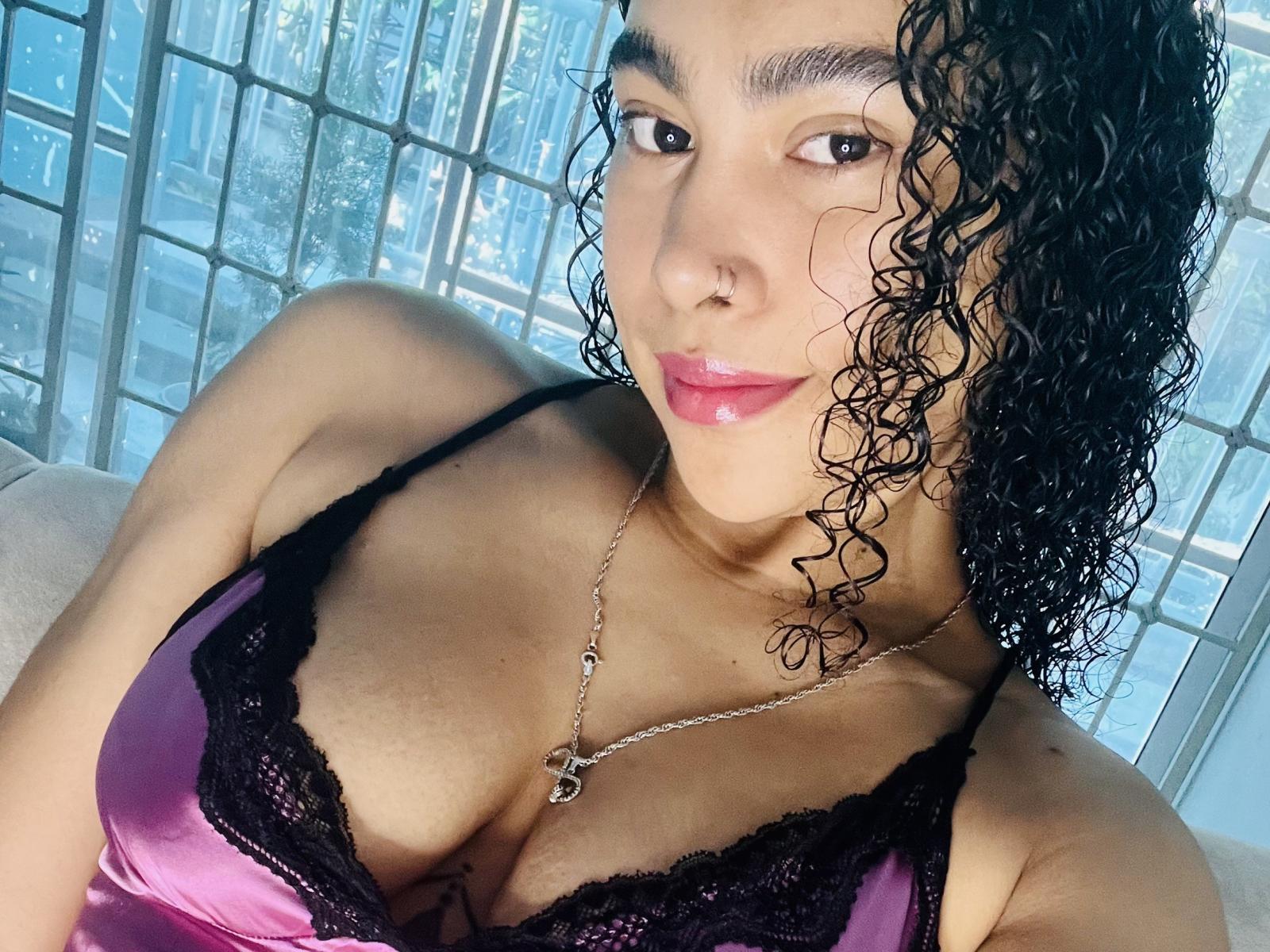 Close-up van SophieDi, een mollige vrouw met donker krullend haar, die een roze en zwarte kanten top met een lage hals draagt, waardoor veel huid te zien is. Een decolleté en een zilveren ketting met een kruisbedel. Ze heeft een verleidelijke uitdrukking..