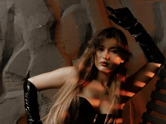 Fotografie von AnneTLuis mit langen, glatten, braunen Haaren, die schwarze Latexhandschuhe und ein schwarzes Top ohne Träger trägt. Sie posiert verführerisch mit erhobenen Armen, im Hintergrund Schatten, intensives Licht, eine sexy Atmosphäre..