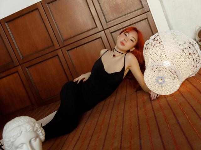 AnnaReyla, una mujer pelirroja atractiva, está acostada en un suelo de madera, vestida con un vestido negro, con las piernas separadas, y tiene una lámpara blanca cubierta de encaje. a la derecha. Ella lleva calcetines blancos. Increíblemente sexy..