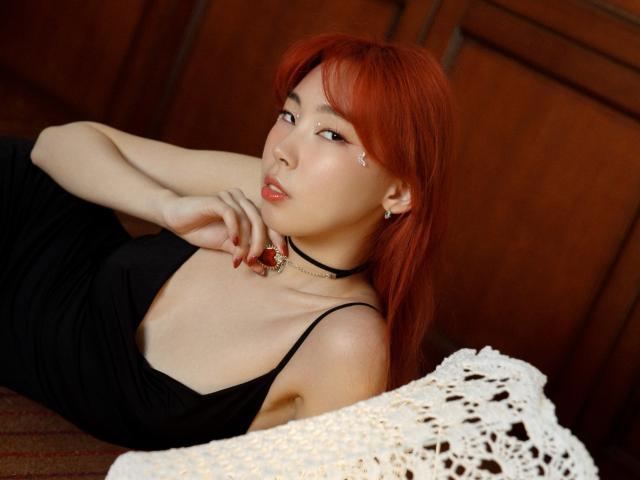 AnnaReyla, avec des cheveux roux, portant une robe noire, allongée sur une surface en bois, une main sur le cou, regardant séductrice l'appareil photo. Dentelle blanche au premier plan, collier ras du cou avec un pendentif rouge..