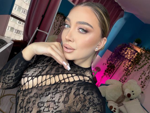 JessicaClarks has light skin and blue eyes, with her blonde hair slicked back. She wears a black, lace, long-sleeve top with a netted pattern revealing her cleavage. Her right hand, with long, white-painted nails, touches her chin. She has pink, glossy lips and subtle, shimmering eyeshadow. She's in a room with pink curtains, a white stand, a stuffed teddy bear, and a lamp with green leaves. The image is a close-up, focusing on her face and upper body.