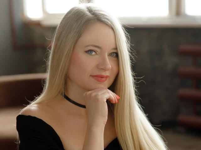 Fotografie von Charlotte Kerti, blonde Haare, blaue Augen, roter Lippenstift, schwarzer Choker, schwarzes Oberteil, Hand am Kinn, weiches Licht. verführerischer, koketter Gesichtsausdruck, Licht von einem Fenster, unscharfer Hintergrund, leichter Schatten im Gesicht..