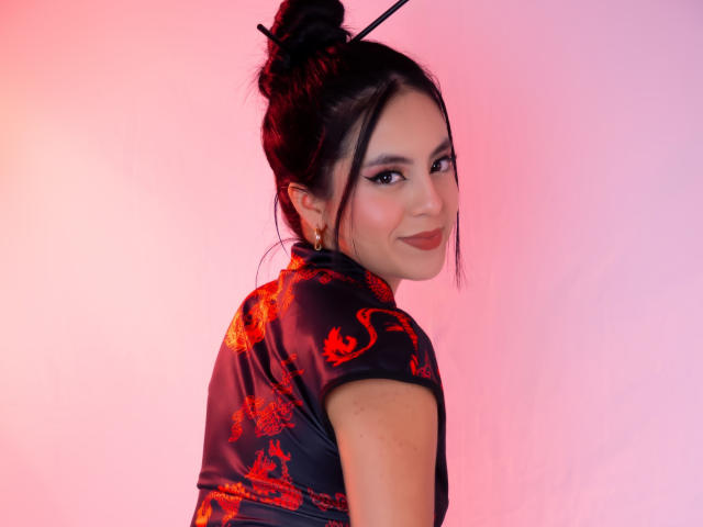 EimmyPark, mit dunklem Haar zu einem Dutt gebunden, trägt ein schwarzes Cheongsam-Kleid mit roten Drachenmustern. Sie blickt mit einem verschmitzten Lächeln zurück, mit einem rosa Farbverlauf im Hintergrund. Fokussiert, sexy, selbstbewusste Pose..