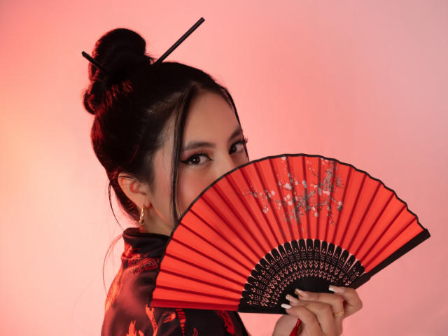 EimmyPark sosteniendo un abanico rojo plegable con detalles negros y brillantes, que cubre parcialmente su rostro. Cabello negro recogido en un moño, fondo naranja-rosa. Las manos agarrando un abanico sugieren una seducción sutil..