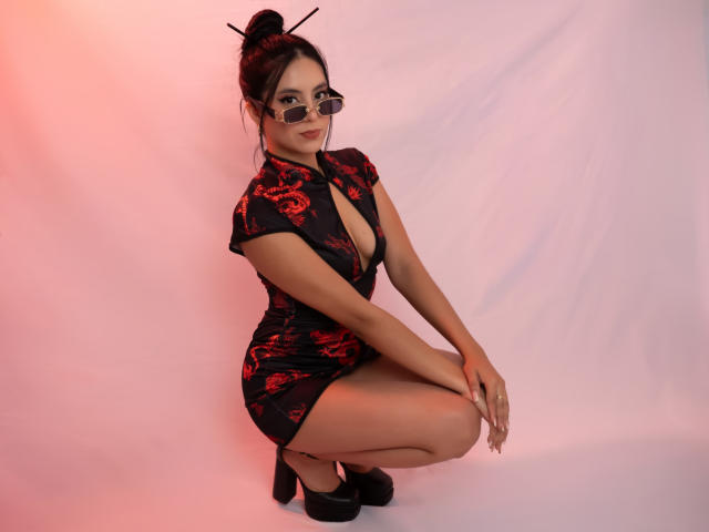 EimmyPark se met en position de squat de manière provocante, portant une robe noire et rouge en dentelle moulante avec un décolleté plongeant, des talons noirs et des lunettes. Ses cheveux noirs sont attachés en un chignon avec des épingles à cheveux. Éclairage rose..