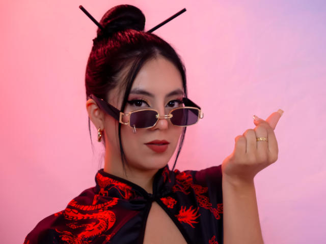 EimmyPark, uma mulher asiática atraente com cabelo preto preso em um coque, vestindo roupas pretas com estampa de dragão vermelho, está segurando... óculos de sol com a armação dourada na mão direita, fazendo um gesto de arma com o dedo..