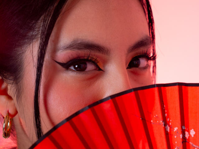 Nahaufnahme von EimmyParks Gesicht mit intensiven Augen, schwarzem Eyeliner und roten Lippen. Sie hält einen leuchtend roten, halbtransparenten Fächer, der ihren Mund teilweise bedeckt. Goldene Ohrringe sind an ihrem Ohr sichtbar..