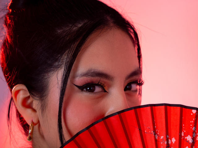 Close-up of EimmyPark, half-visible face with dark hair, bold eyeliner, holding a red, translucent fan in front of her mouth. Pink and red background, gold earrings, intense gaze.