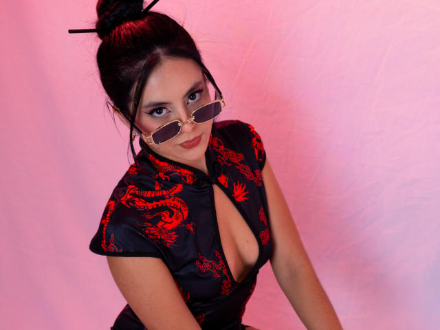 EimmyPark, met donker haar in een knot, draagt een zwarte jurk met rode drakenmotieven en een diepe decolleté, en ronde zonnebril, voorovergebogen met een verleidelijke blik, roze achtergrond.