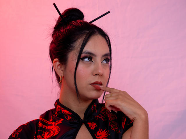 EimmyPark, mulher asiática com cabelo preto preso em um coque, blusa com estampa vermelha e preta, olhar sedutor, mordendo o dedo, lábios vermelhos. Fundo com gradiente rosa e branco, dois prendedores de cabelo, clima sensual..