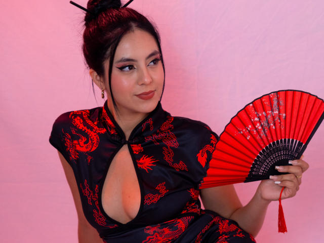 EimmyPark, una donna asiatica con i capelli scuri raccolti in uno chignon, indossa un cheongsam nero con motivi floreali rossi, e tiene in mano qualcosa di rosso. Ventaglio pieghevole con dettagli neri. Sfondo rosa, espressione seducente
.