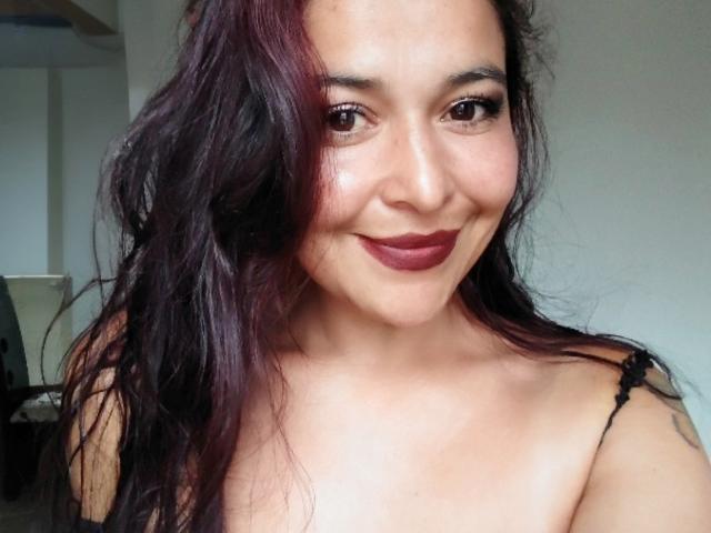 Nahaufnahme von AfroditaLaDiosa, die lächelt, mit dunkelrotem Lippenstift, langen schwarzen Haaren, heller Haut und einem schwarzen Top mit Trägern. Sichtbarer Ausschnitt, leichte Schattierung auf der rechten Wange, lockeres Foto..