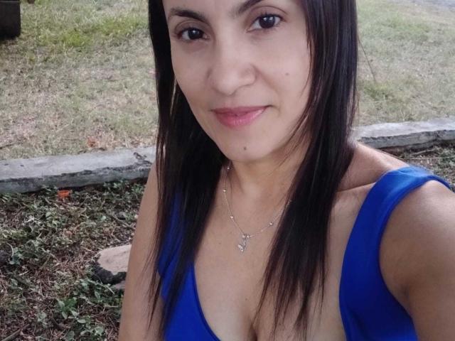 Photograph of SolaraDubois with long black hair, wearing a blue tank top, showing cleavage, smiling subtly, wearing a delicate necklace. Grass and concrete edge in the background. Fucking cute.