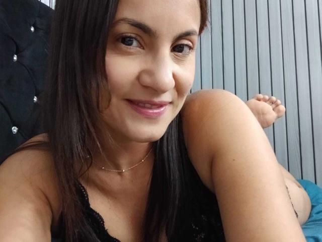 Close-up selfie of SolaraDubois with dark brown hair, smiling, wearing a black lace top, showing one raised leg. She's wearing a thin gold necklace, looking directly at the camera. Fucking sexy.