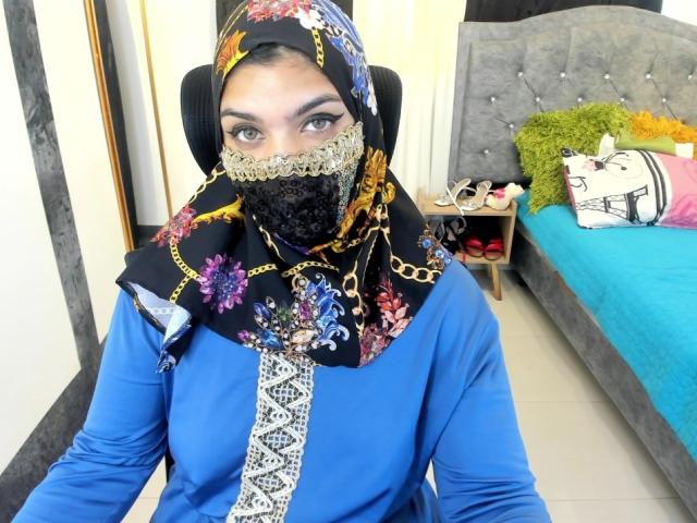 Aniisa Ali, con la pelle di un marrone medio, indossa un hijab nero con fiori, una camicia blu a maniche lunghe e una mascherina di pizzo nero, ed è seduta. Vicino a un divano blu con cuscini verdi e gialli. Tensione sessuale evidente..