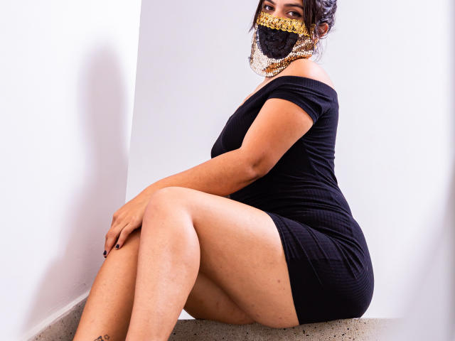 Aniisa Ali, sentada em um canto branco, vestindo um vestido preto com um ombro descoberto e uma máscara com padrão dourado, com as pernas cruzadas, exibindo suas coxas definidas. Ela está olhando de forma sedutora, com um toque de atração..