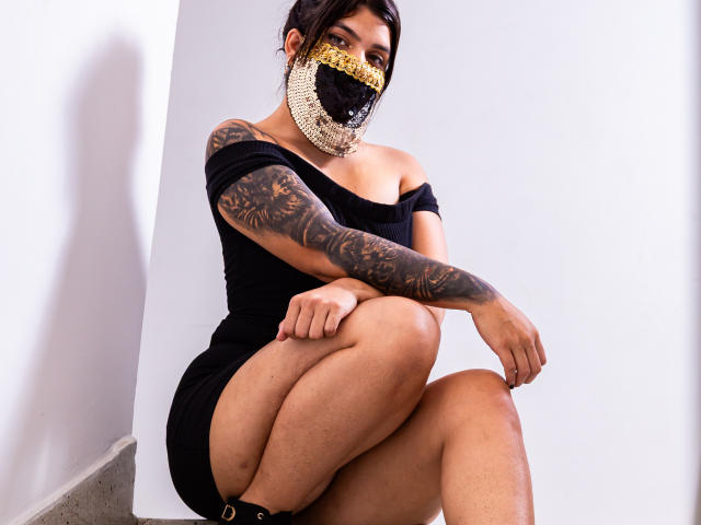 AniisaAli est assise, les jambes écartées, vêtue d'une robe noire à une épaule et portant un masque doré. Elle a des tatouages sur les bras et une silhouette voluptueuse, dégageant une forte sensualité. Fond blanc éclatant.