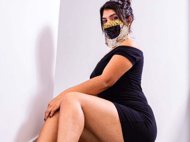 AniisaAli, assise, les jambes croisées, porte une robe noire à une épaule et un masque en dentelle noire orné de motifs dorés. Elle regarde de côté avec une expression séduisante et intense. Fond blanc.