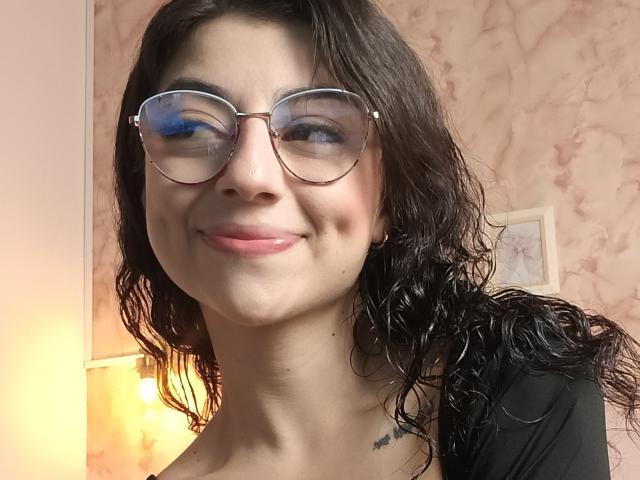 Fotografia di Anntonella Watson, sorridente, che indossa occhiali rotondi grandi, capelli neri ondulati, top nero, pelle chiara, aspetto delicato. trucco, luce gialla tenue proveniente da una lampada sullo sfondo, tatuaggio discreto sulla spalla destra..