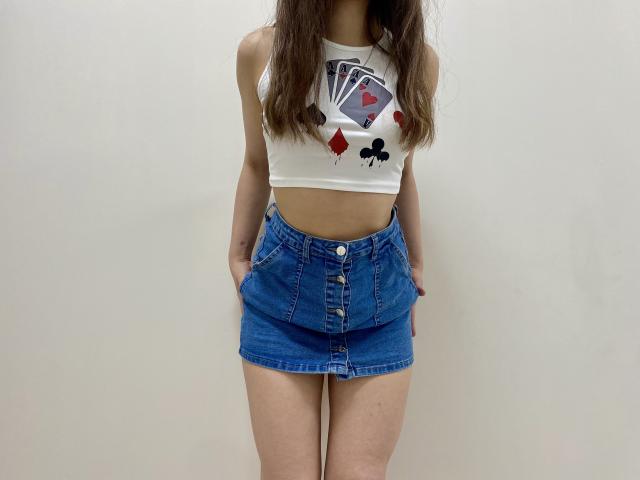 JoannaSia stands facing forward, wearing a white crop top with a playing card design and red diamonds, and high-waisted blue denim skirt. Her hands rest in her pockets. Tight, casual, sexy.