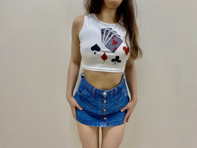 JoannaSia met lang bruin haar, gekleed in een witte crop top met kaartmotieven en een blauwe denim mini-rok, handen in haar zakken, staand tegen een effen witte muur. Heel aantrekkelijk..
