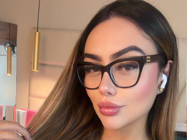 Close-up of MissAry, a young woman with long brown hair, black-framed glasses, and full pink lips, wearing white earrings. She's gazing directly at the camera with a slight smirk. Background includes a modern interior with hanging lights.
