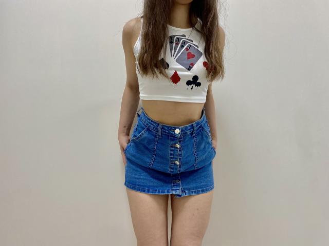 Joanna, con lunghi capelli castani, indossa un top corto bianco con una grafica e una gonna a vita alta di jeans blu. Mani nelle tasche, in piedi con le gambe leggermente divaricate. Sfondo bianco..