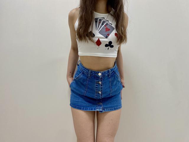 JoannaSia está de pie, con las manos en una falda vaquera azul de cintura alta, una camiseta blanca corta con un diseño de trajes de cartas, y cabello castaño largo. Fondo blanco, ligera tensión sexual, pose casual, complexión delgada..