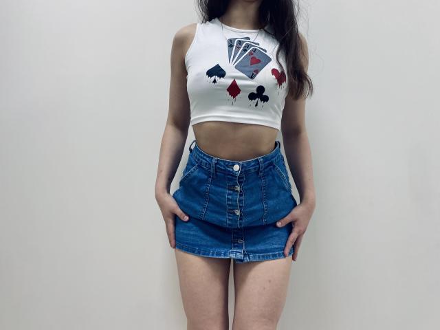 JoannaSia draagt een witte crop top met kaartmotieven en een blauwe spijkerrok, met haar handen in de zakken. Haar buik is zichtbaar, wat een sexy en zelfverzekerde houding benadrukt. Strakke rok.