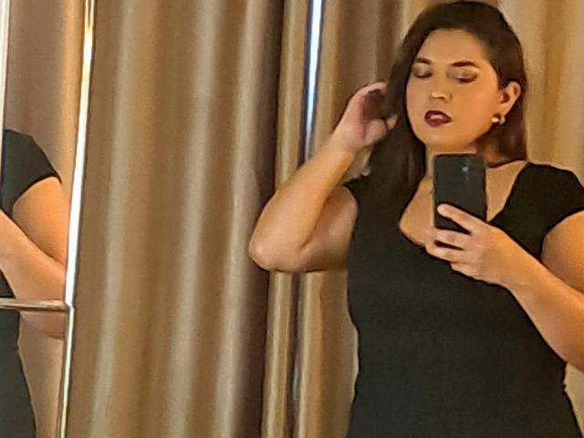 Photograph of PaulaAnd taking a mirror selfie. She's wearing a black dress, has dark brown hair, and red lipstick. Her left hand touches her hair, right hand holds a phone. Reflects her side profile.
