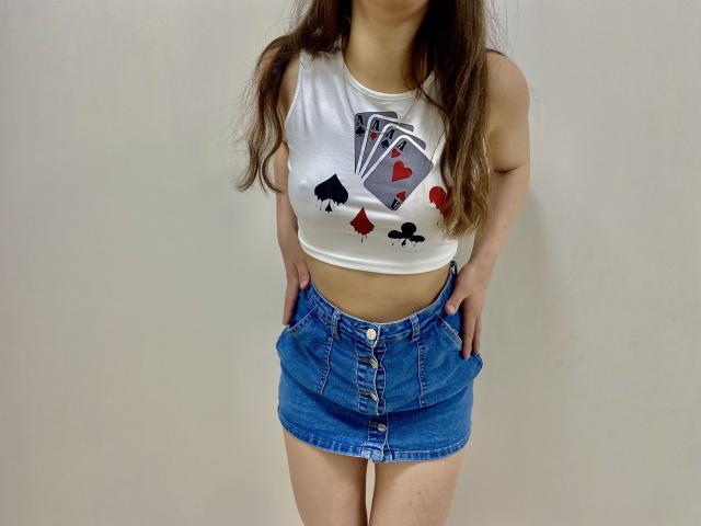 JoannaSia, magra, vestindo uma blusa cropped branca com estampa de cartas de baralho e uma saia jeans azul, com as mãos nos bolsos, cabelos castanhos longos. cabelo, fundo branco. Muito atraente, pose confiante, estilo casual..