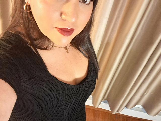 Close-up of PaulaAnd, a fair-skinned woman with dark brown hair, wearing a black textured dress, red lipstick, and gold hoop earrings. Golden curtains in the background. Fucking sexy.