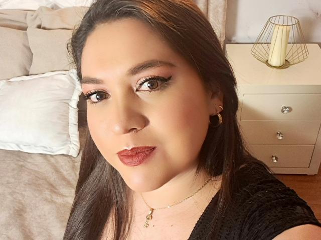 Primo piano di PaulaAnd, una donna latina con lunghi capelli neri, rossetto rosso e eyeliner scuro, che indossa una maglietta nera, in un camera da letto con una testiera beige e una lampada bianca. Incredibilmente attraente..